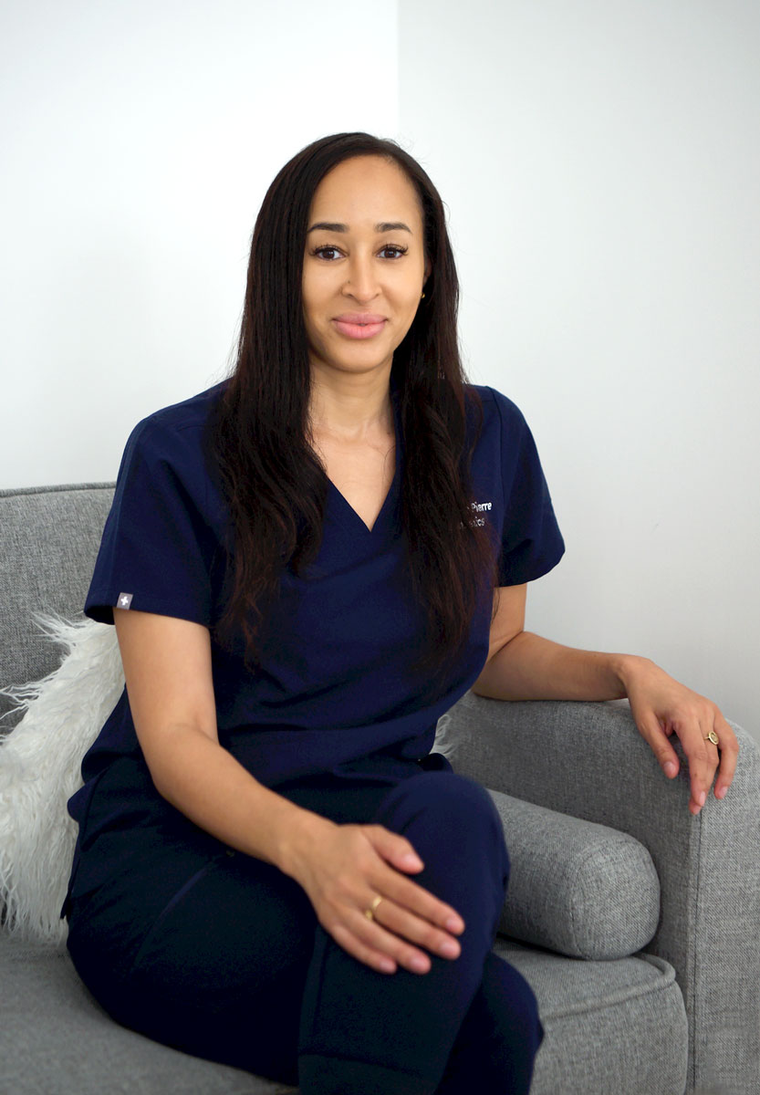 Dr-Louise-Pierre-Sofa-Portrait Aesthetic practitioner seated on a sofa in a clinic setting, wearing navy scrubs.