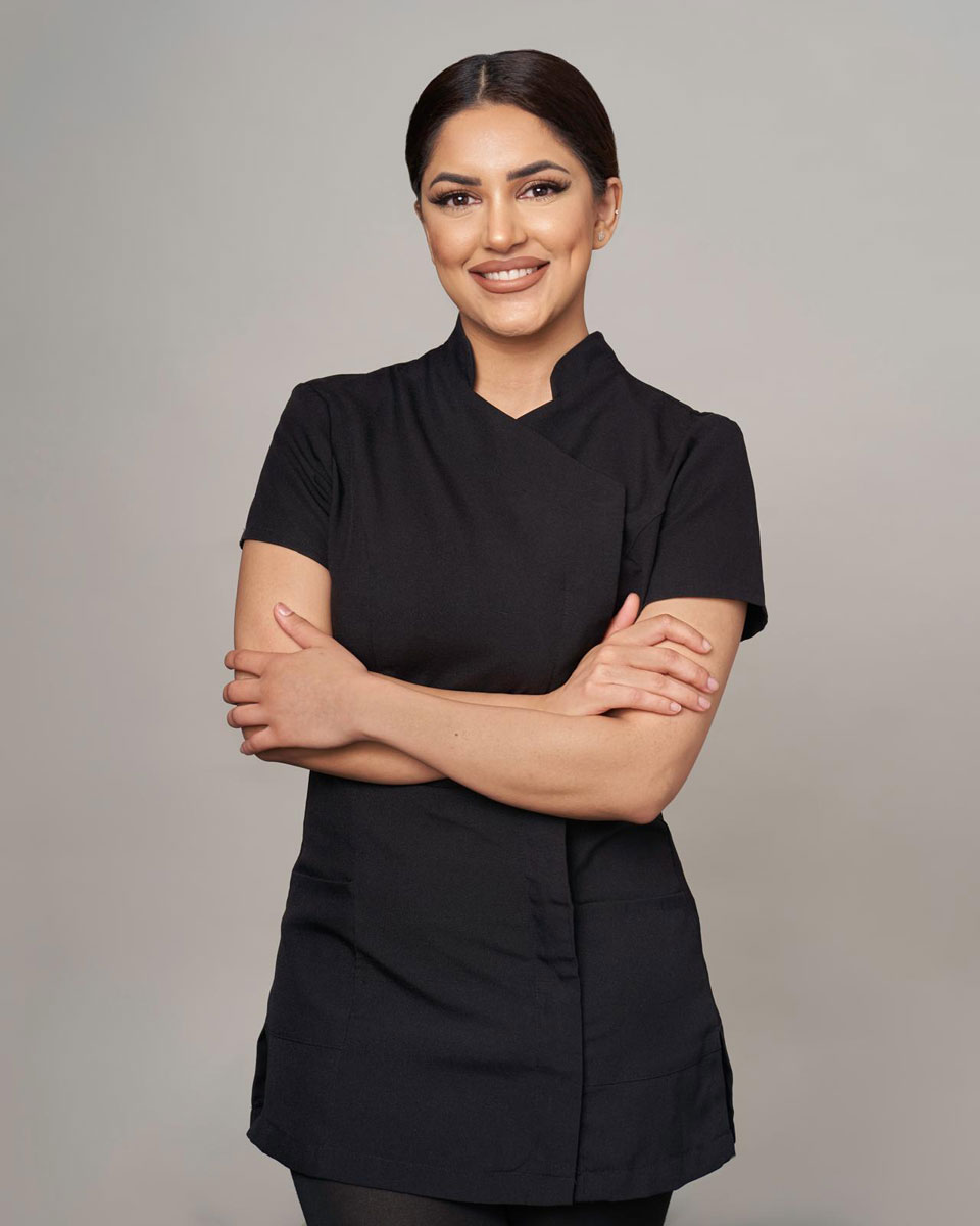 Aesthetic practitioner smiling with arms crossed in a black clinic uniform against a neutral backdrop.
